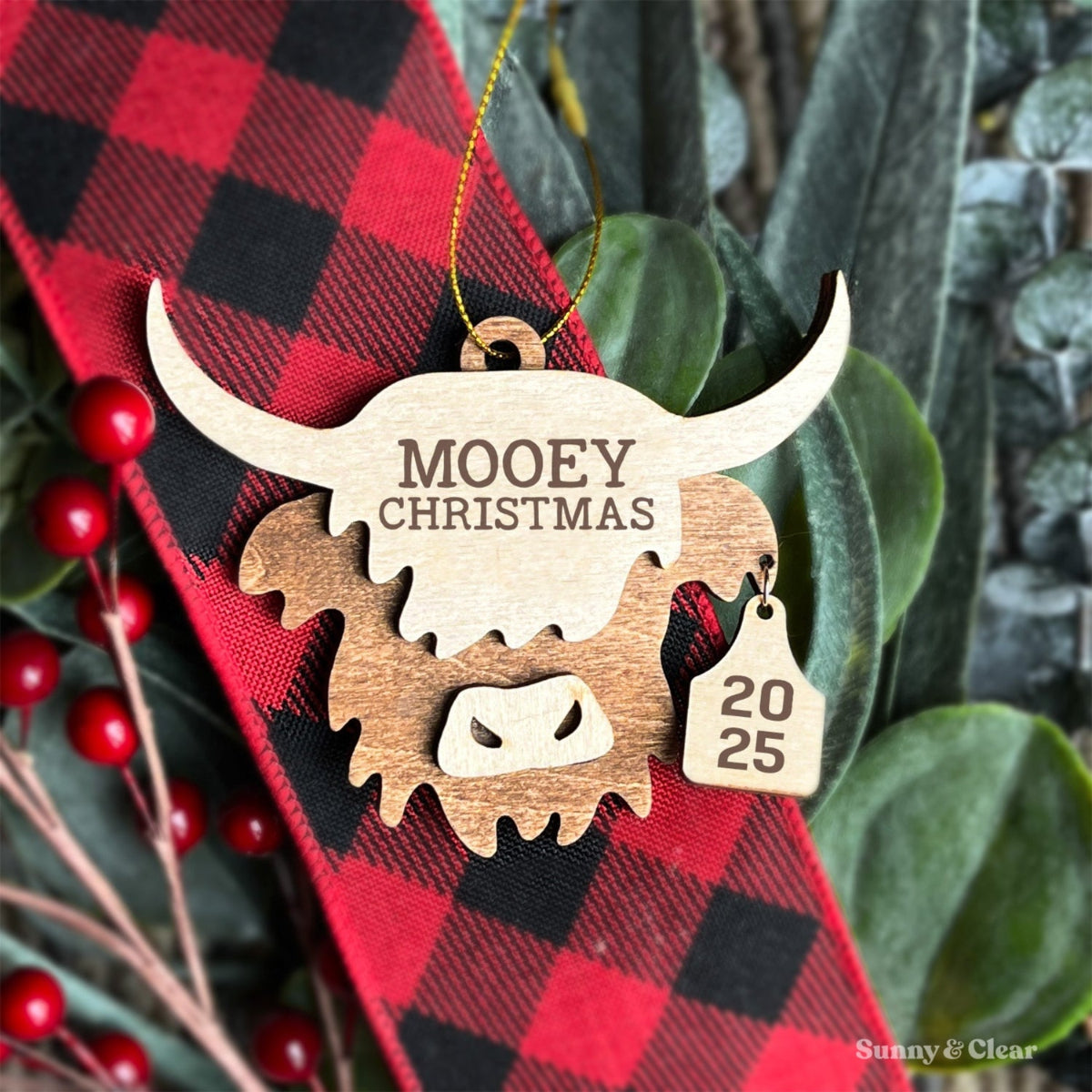 Wooden Christmas ornament with 'MOOey Christmas' and '2025' on a red and black checkered fabric background.