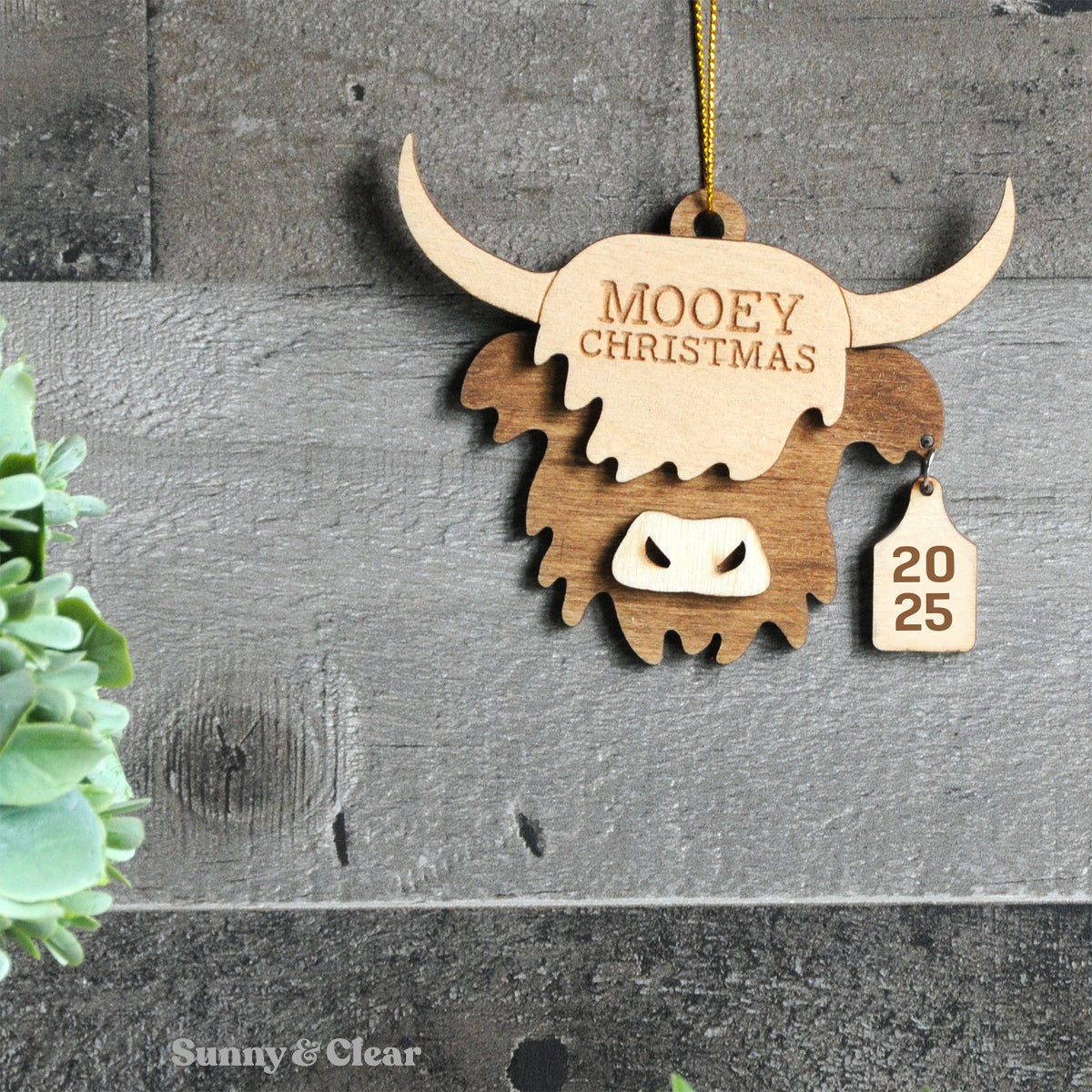 Wooden ornament shaped like a highland cow with 'Mooey Christmas' text on a gray surface.